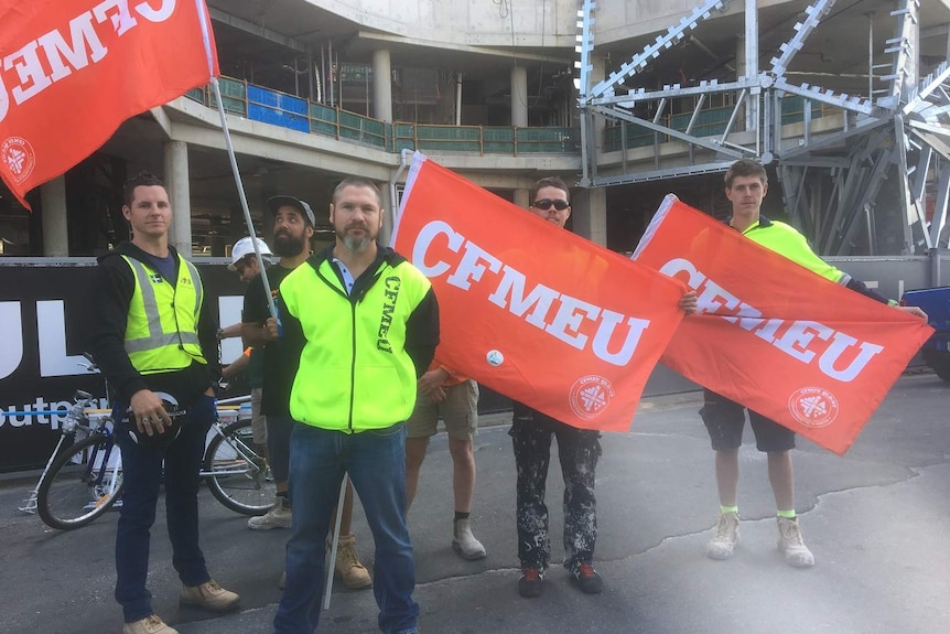 Union members stand with red and white CFMEU flags in front of a building site.