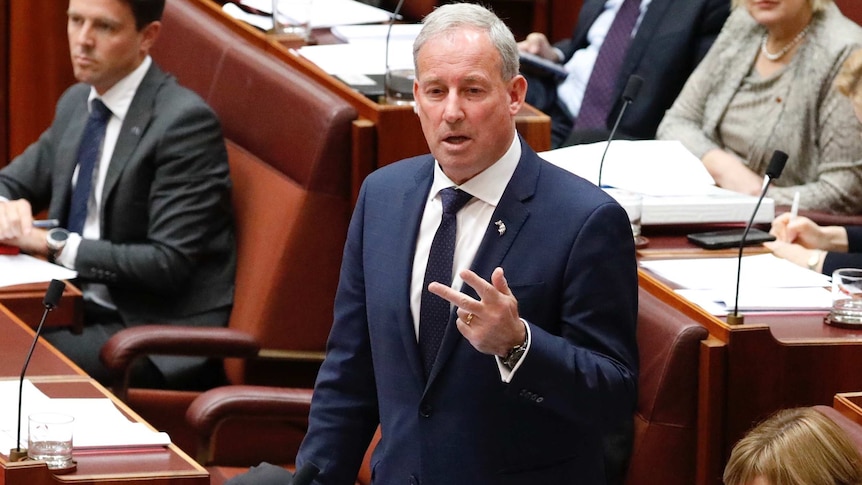 Senator Richard Colbeck stands and talks wearing a dark blue suit, white shirt and blue tie