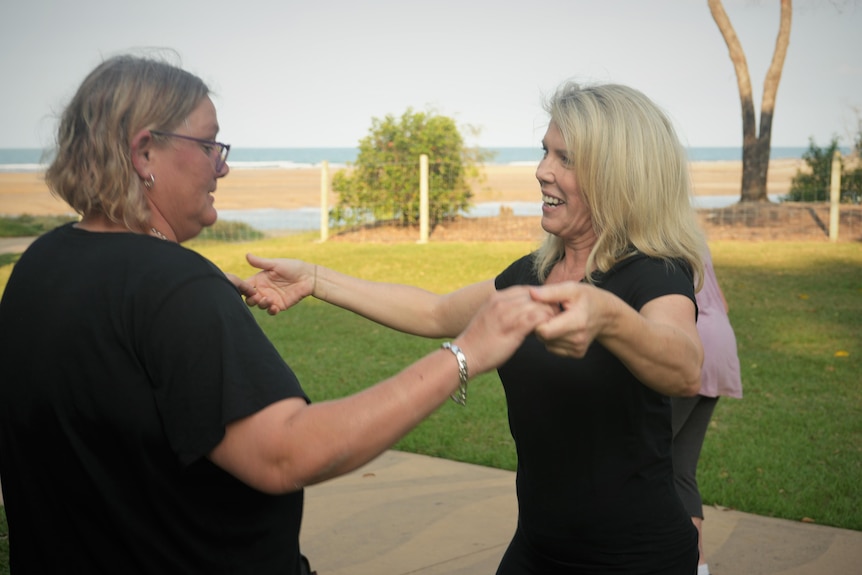 Two women dance in a park, with the beach in the background