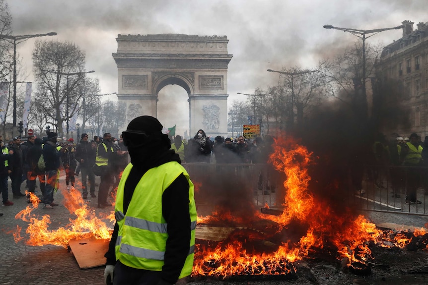 Arc de Triomphe in the background with flames in middle ground and man wearing black mask and yellow hi-vis vest in foreground.