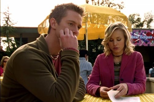 Logan  has his hand on his face as he looks off in the distance next to Veronica, who stares down at paper.