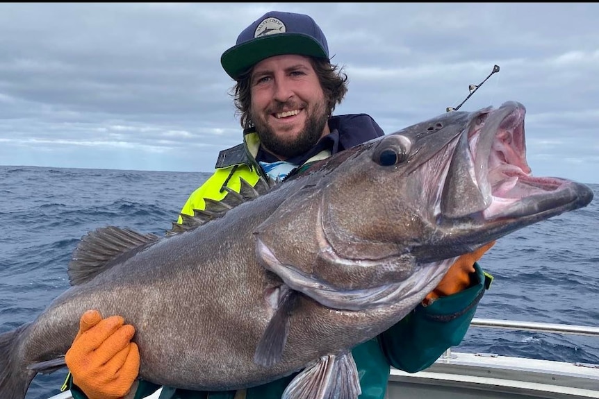 A man in a cap smiles at the camera holding a huge fish, the ocean behind him