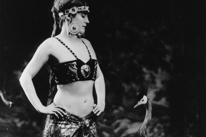 Black and white photo of Annette Kellerman dressed in decorated crop-top, skirt and large earrings, looking down at a peacock.