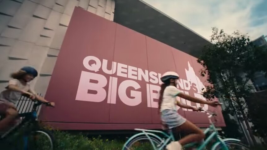 A pair of kids on bikes ride past a large billboard that says "Queensland's Big Build".