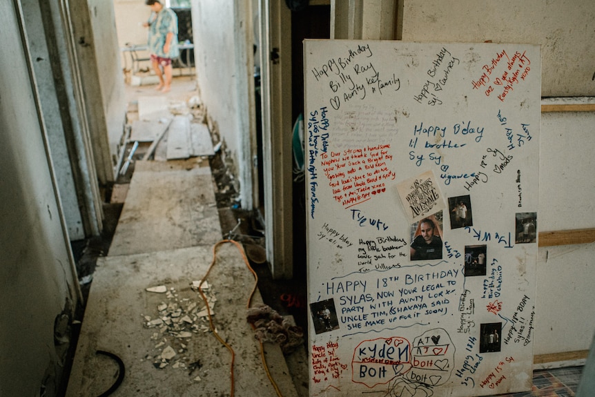 Boards cover holes in the damaged floor of a hallway. A large board with written birthday messages is propped against a wall.