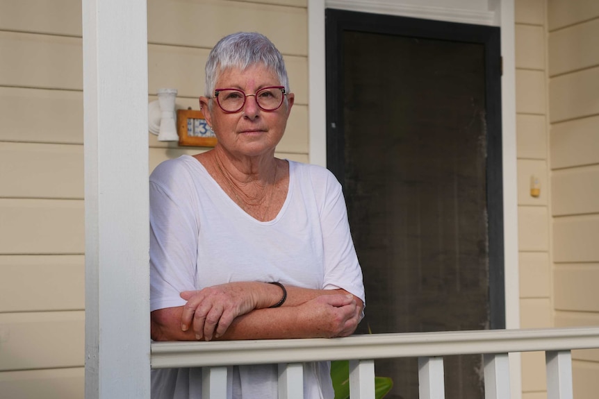 A woman with short grey hair and wearing glasses leans on a verandah railing with a serious look on her face.