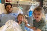 A man lying in a hospital smiling with his two young daughters, also smiling.