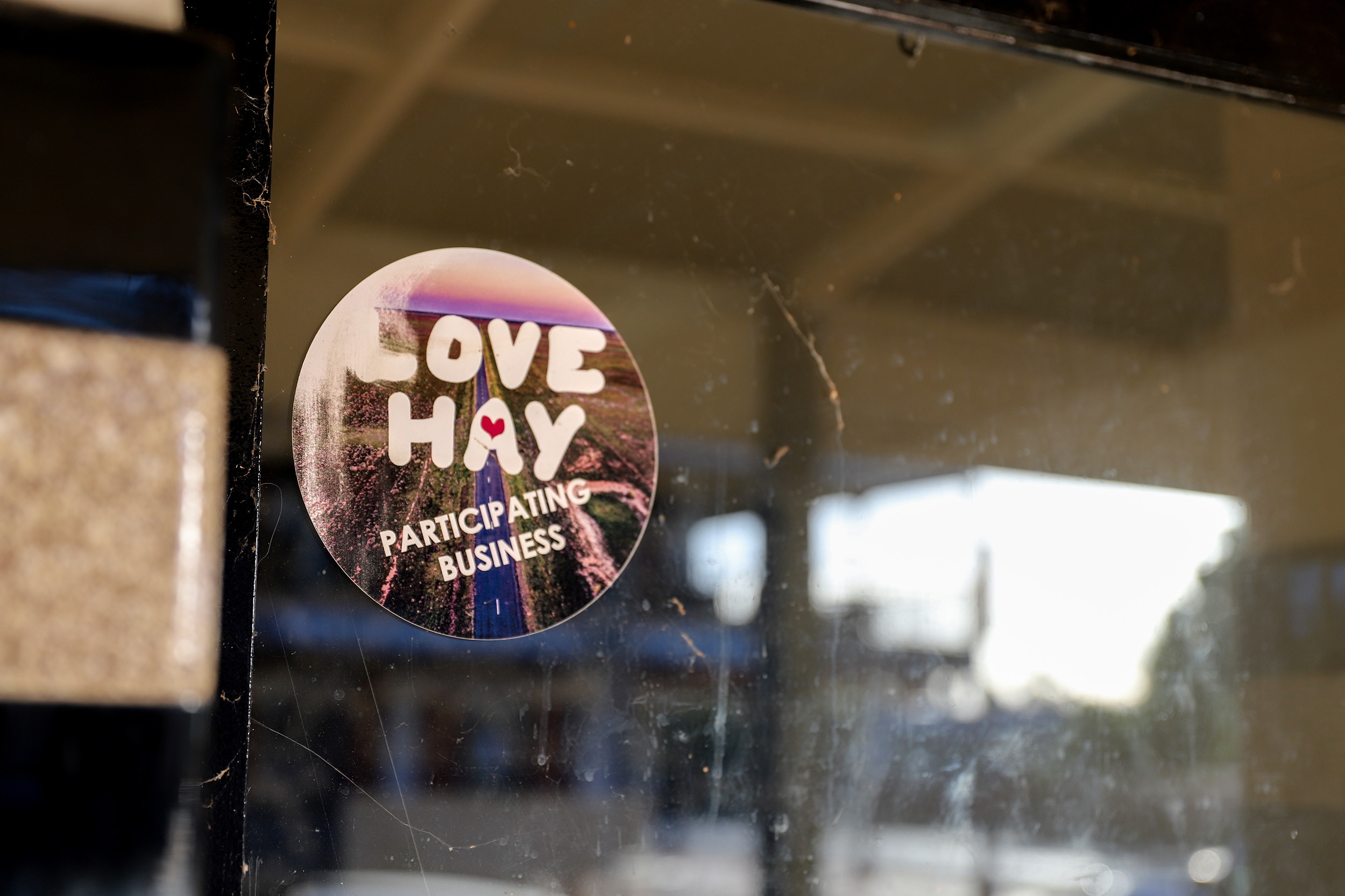 A sticker reading 'Love Hay participating business' stuck on a window with a reflection of a building in the glass.