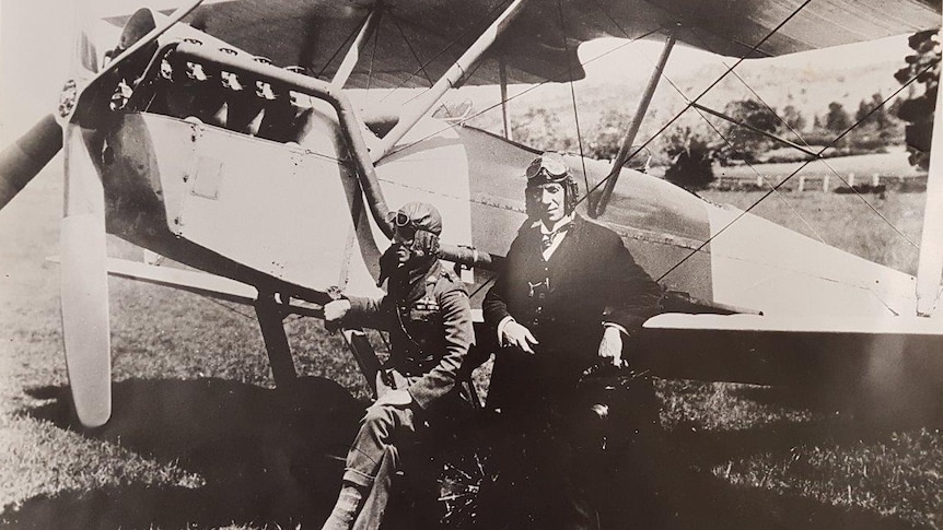 Historic monochrome photo of two men standing by plane in a field, wearing aviator headgear and goggles.