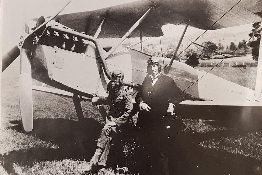 Historic monochrome photo of two men standing by plane in a field, wearing aviator headgear and goggles.