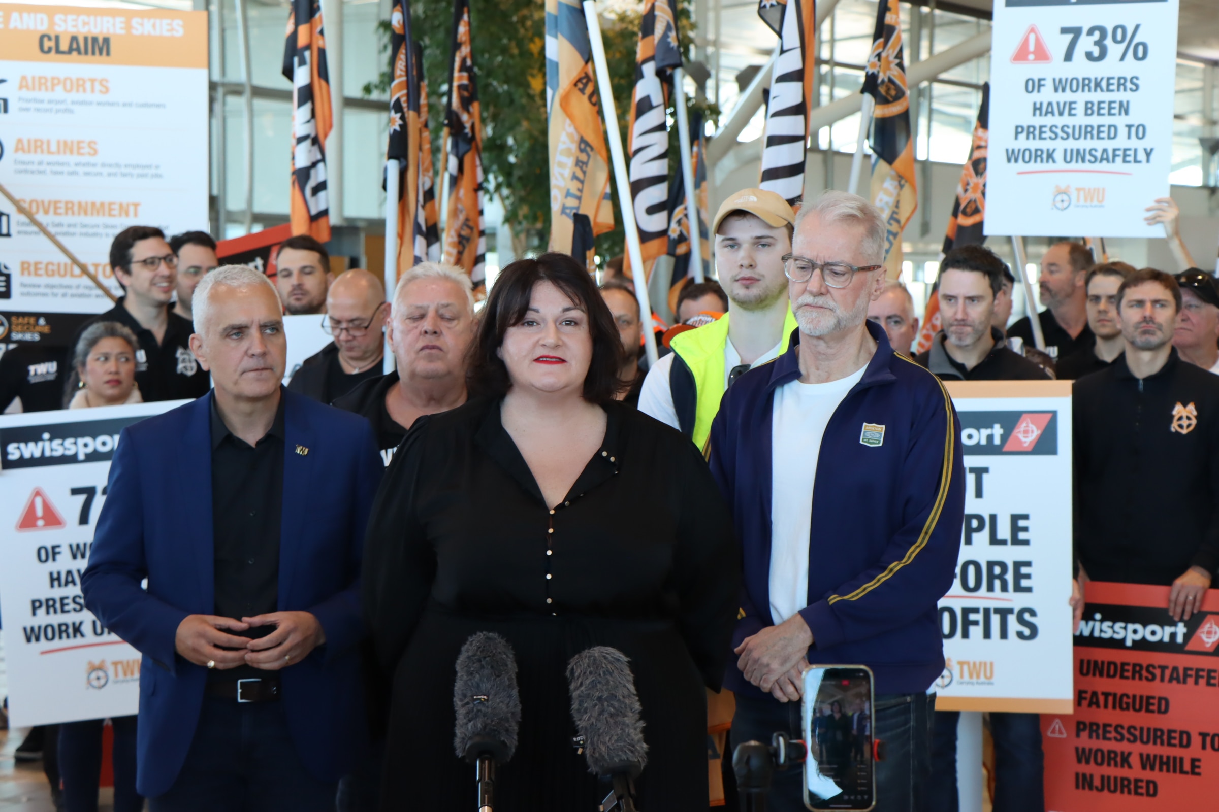 a woman in a black dress standing in front of a crowd of union members speaking to microphones