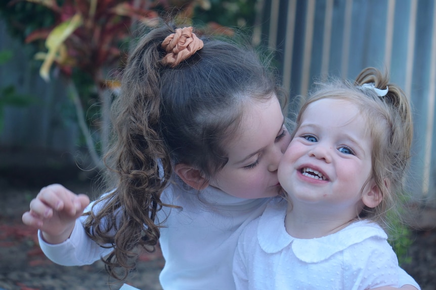 Arlo, petite fille aux longs cheveux bruns, embrassant sa petite soeur Banks, petite fille aux yeux bleu vif, cheveux blonds.