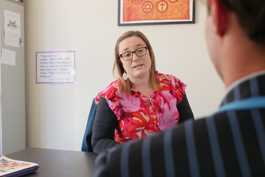 A woman in glasses and a pink top speaking to a person in an office.