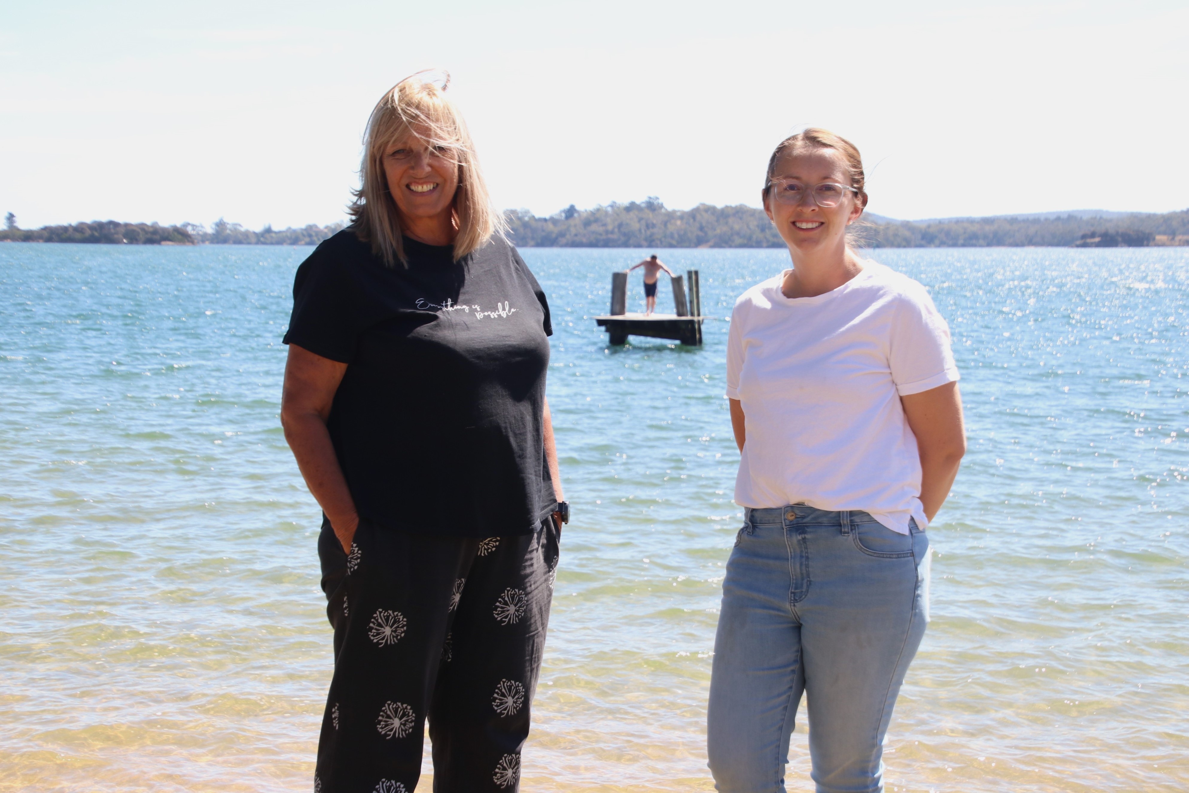 Two women at the beach stand in front of a free standing diving board out at sea.