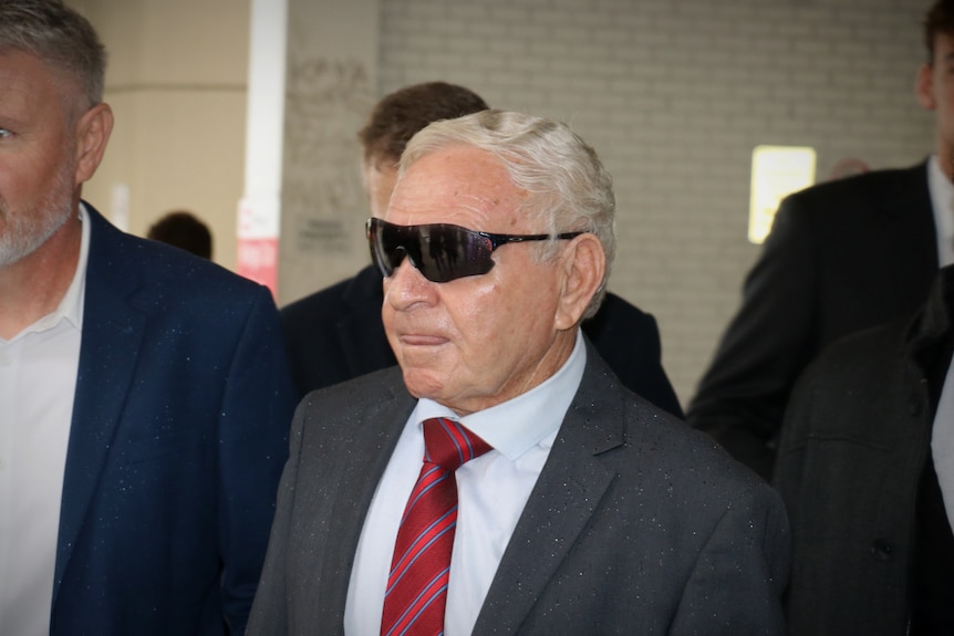 A man with sunglasses and a red tie walks outside of a courthouse