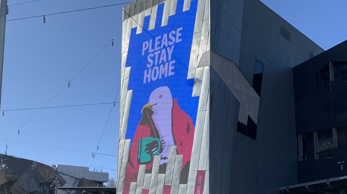 A sign reads 'Please Stay Home' with artwork of a seagull wrapped in a blanket holding a mug.