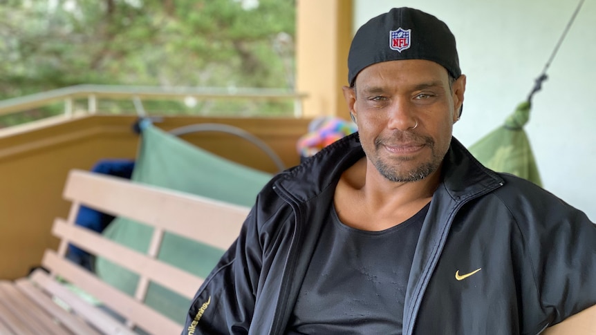 A man smiles at the camera wearing a black cap backwards while sitting on a bench