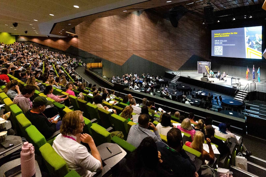 Hundreds of people sit in a large auditorium, looking towards an elevated stage where four people are having a panel discussion.