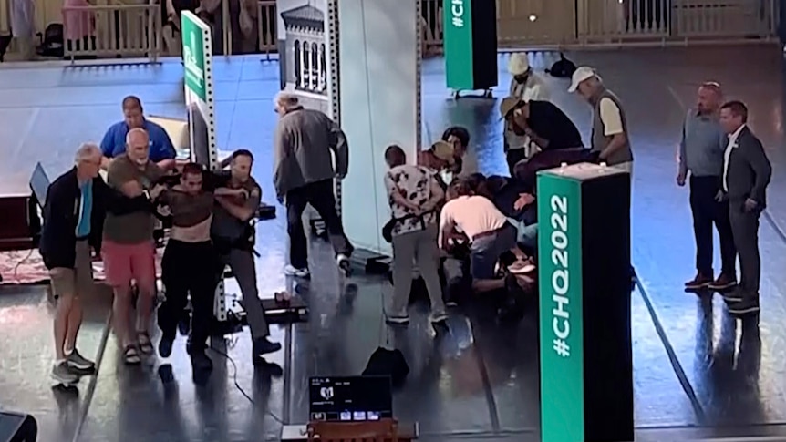 A grainy video still shows a group of people around a person on the floor, as three other people lead another man away