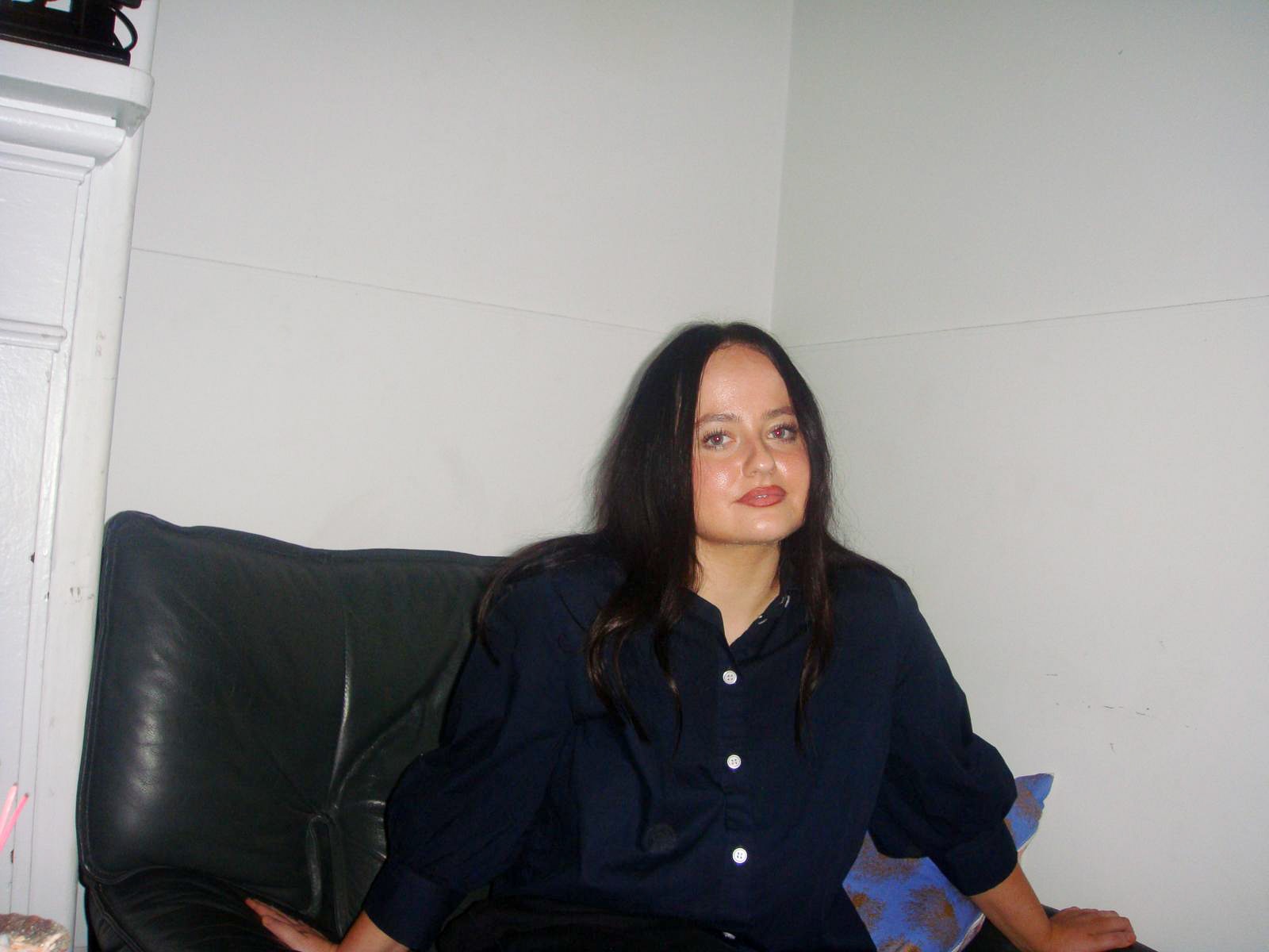 A young woman with brown hair sits on a couch, smiling