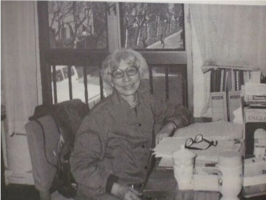 A woman with grey hair and glasses sits at a desk with a window behind her.