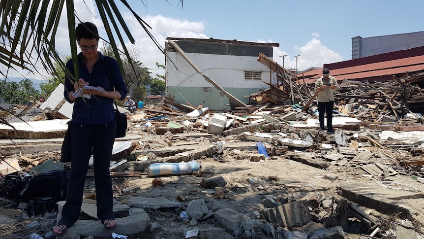 Barker writing on notepad and Hemingway holding drone controls standing amid piles of rubble.