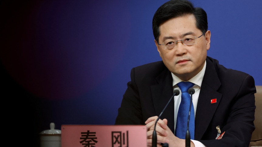 A neatly groomed Chinese man wearing a suit and blue tie sits at a news conference table.