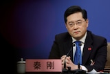 A neatly groomed Chinese man wearing a suit and blue tie sits at a news conference table.