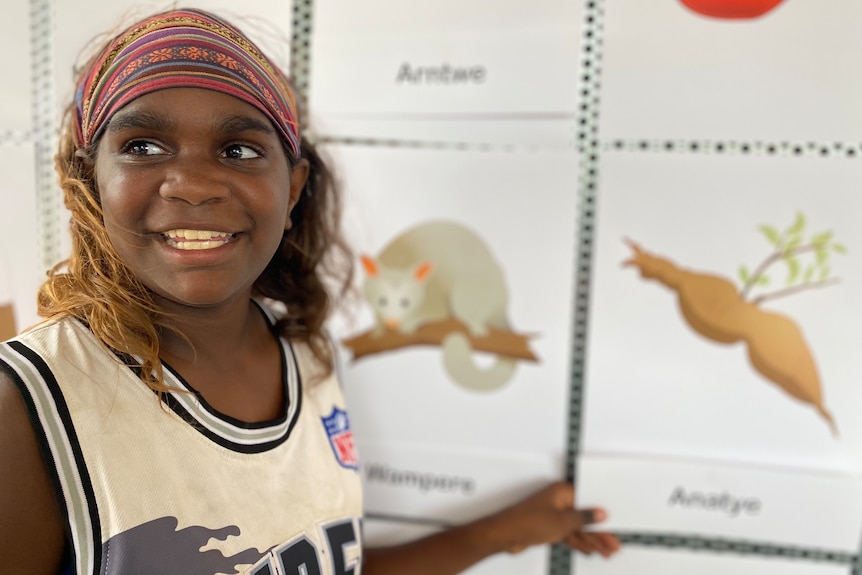 A young child smiles while pointing to emoji's printed out and pinned on the wall behind her.