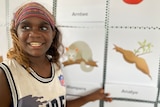 A young child smiles while pointing to emoji's printed out and pinned on the wall behind her.