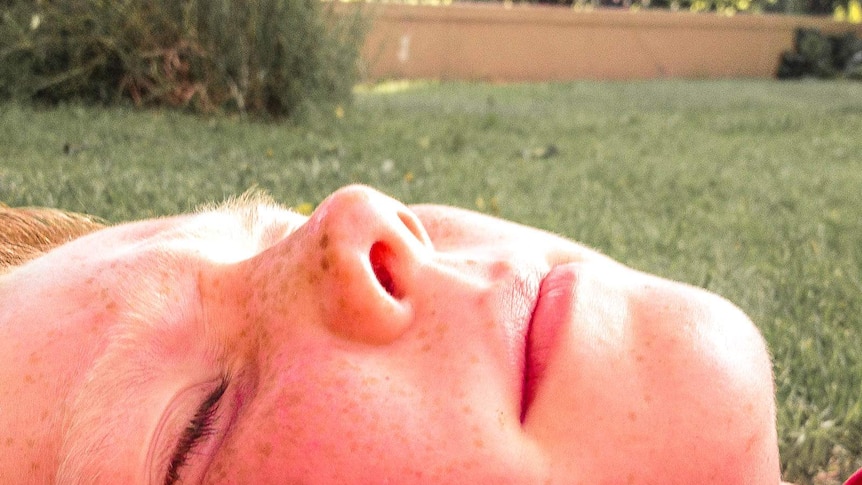 A pink faced boy lying in the sun on grass