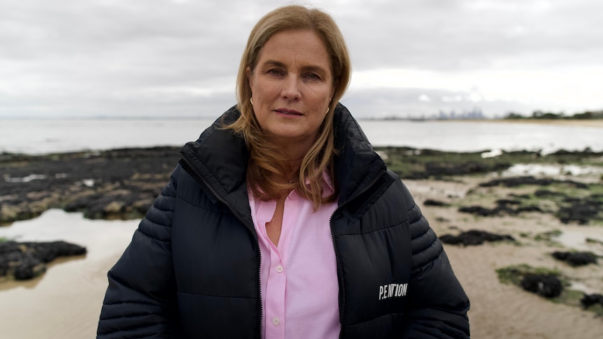 Woman in a pink shirt and black puffer jacket standing on the shore. The sky is overcast.