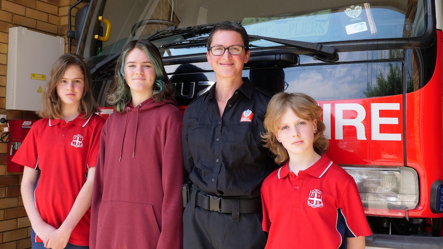 A female fire fighter stands in front of a fire truck with her three daughters by her sides. 