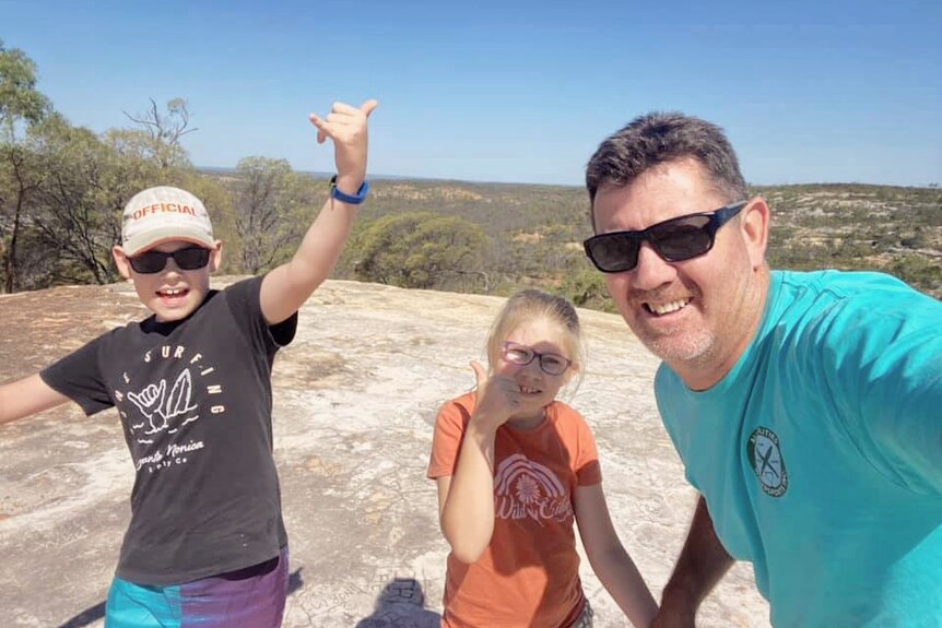 A father wears a green shirt and poses for a photo with his son and daughter atop a cliff on a bushwalk