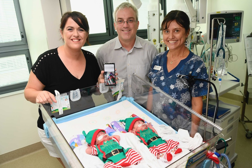 Samantha Hayden holds her phone, displaying sons Zachary and Sebastian, while standing over the crib with two other people.