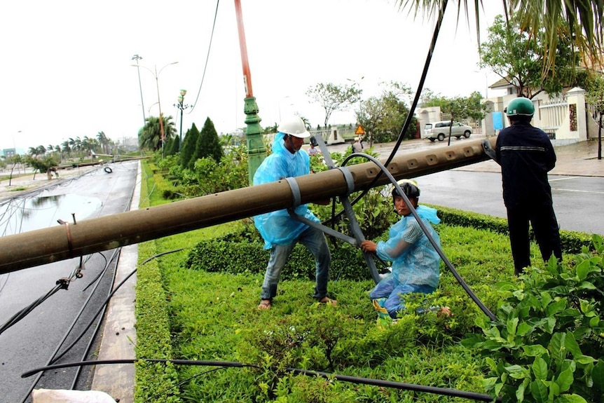 Men wearing helmets repair a fallen electricity pole on a nature strip in the middle of the road.