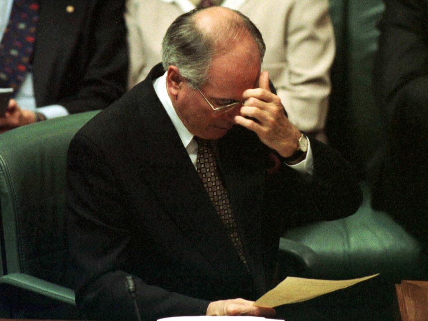 John Howard sits in a green chair, reading a document and wiping his brow.