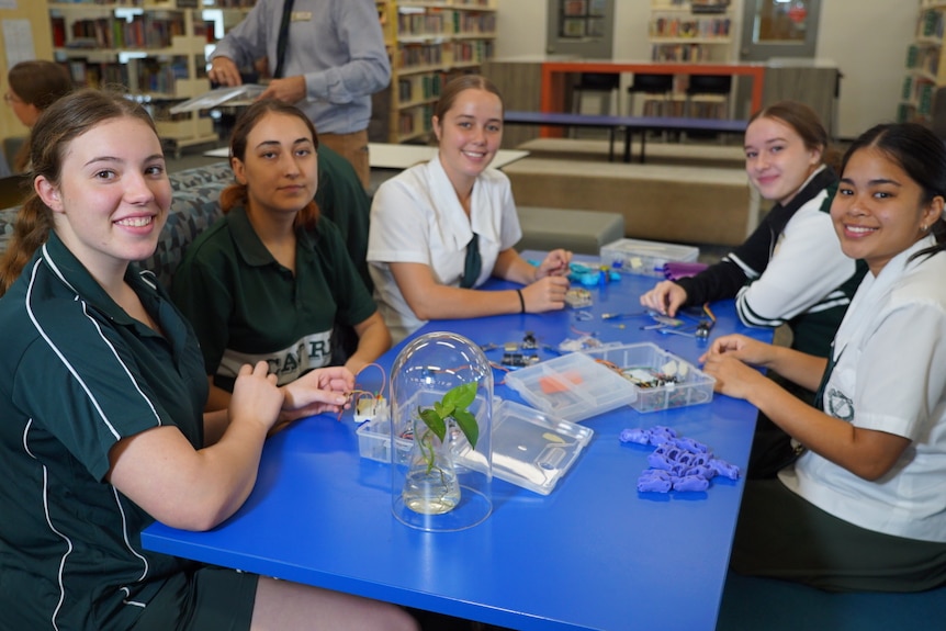 Five female students sit around a blue table working. Their uniforms are white and green. 