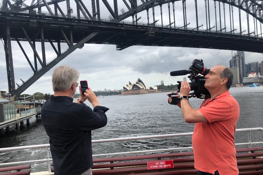 Cameraman filming Neill looking at mobile phone with Opera House and Sydney Harbour Bridge in background.