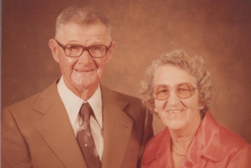 A smiling older couple, both formally dressed and wearing glasses.