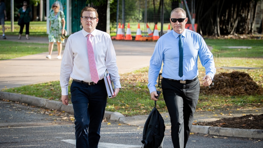 Two serious-looking men walking through a carpark on a sunny day.