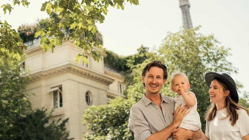 A young man and woman holding a one year old boy with buildings in Paris behind them.