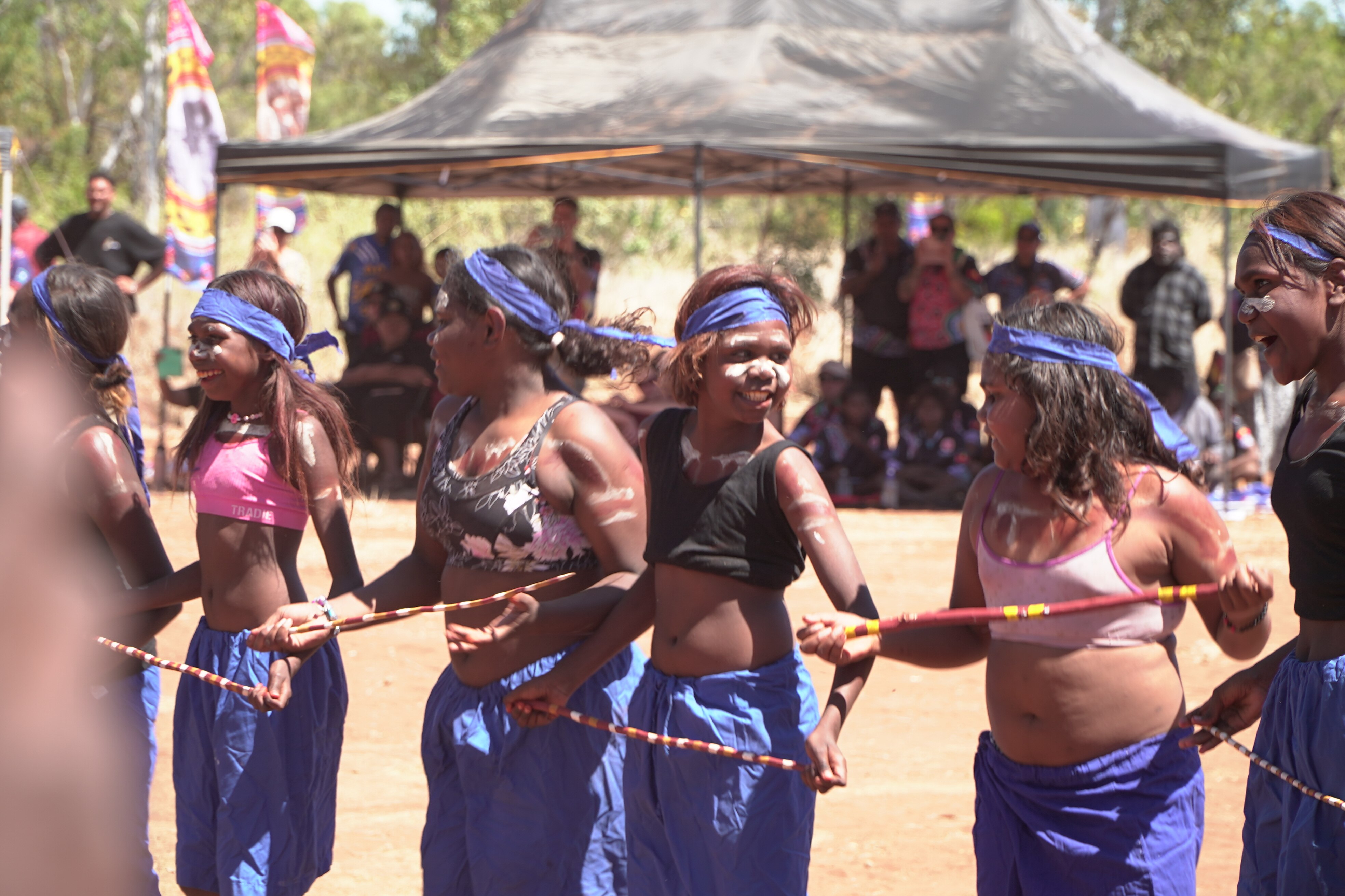 A line of young Aboriginal girls holding red-painted sticks, blue pants, cropped tops, blue headbands. 
