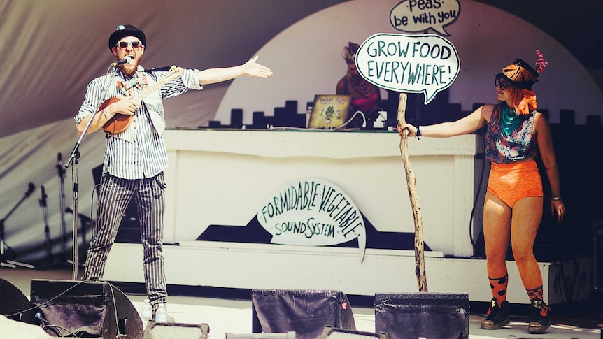 A man and woman in bright costumes singing on stage with signs promoting gardening.