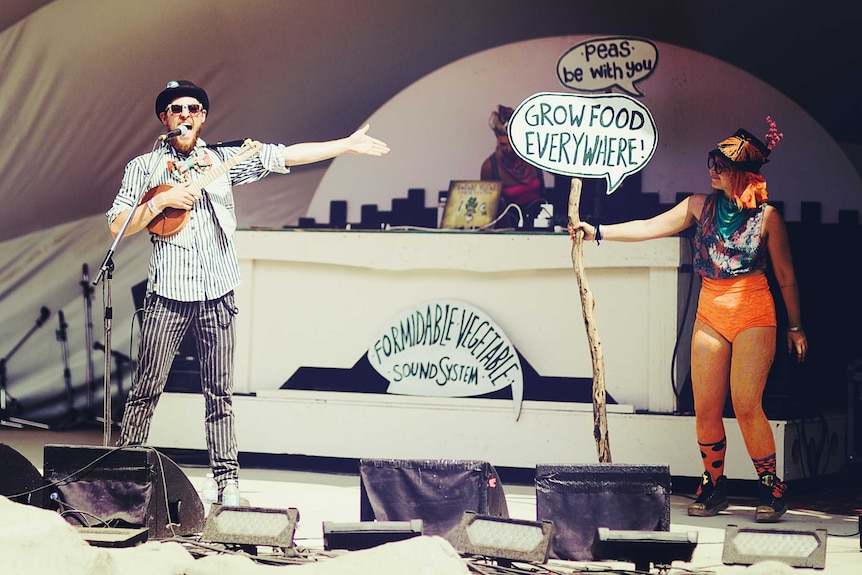 A man and woman in bright costumes singing on stage with signs promoting gardening.