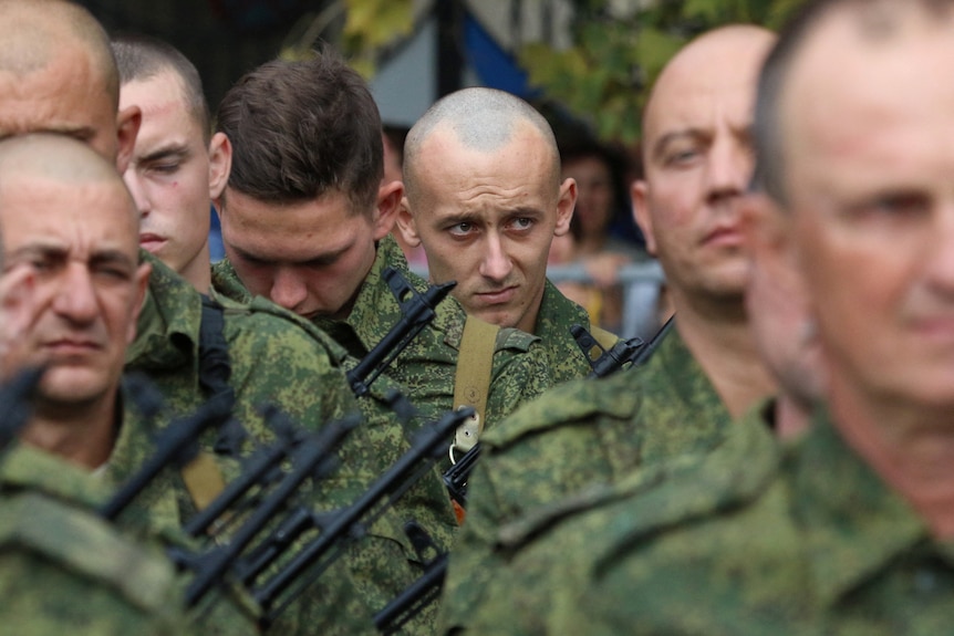 A man with a shaved head stands among other military recruits looking sheepish