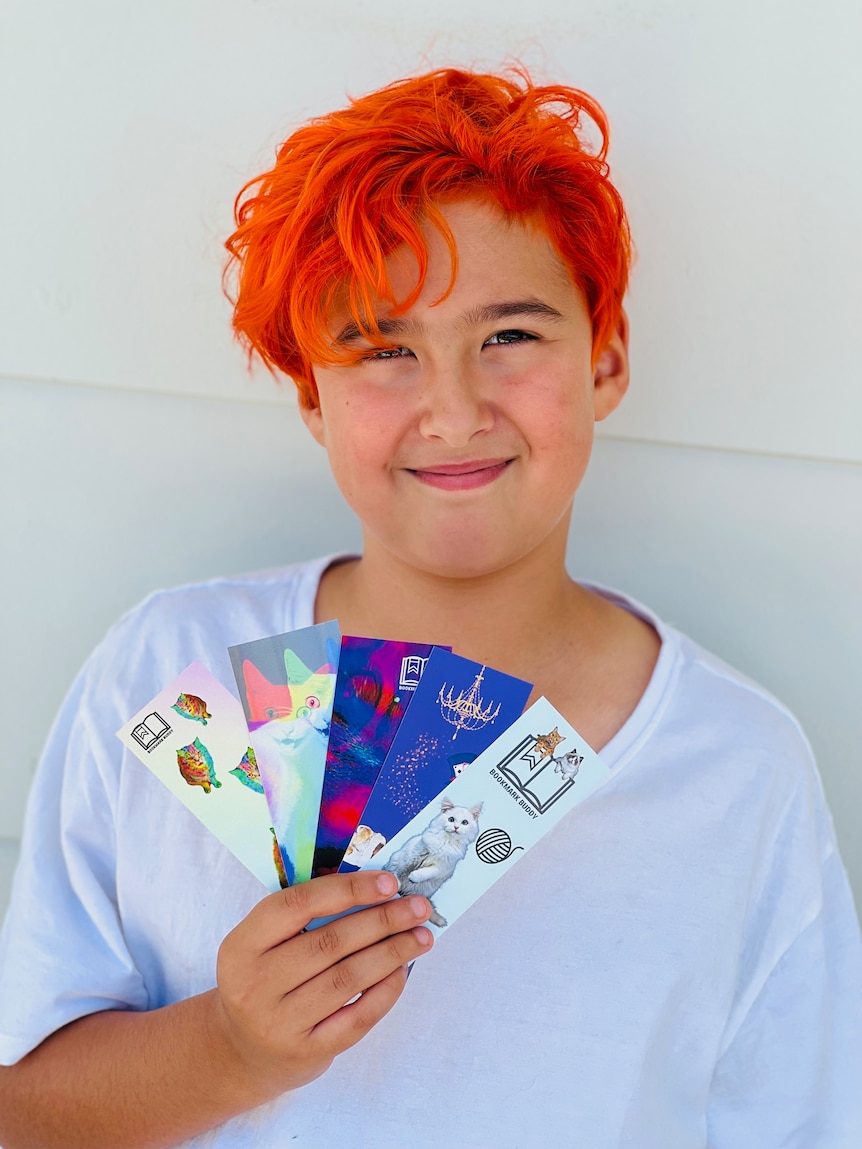 A young boy smiling at the camera while holding colourful bookmarks he designed himself