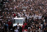 A large crowd of people with signs saying 'Justice pour Nahel' are pictured walking in a march, a white van drives through it.