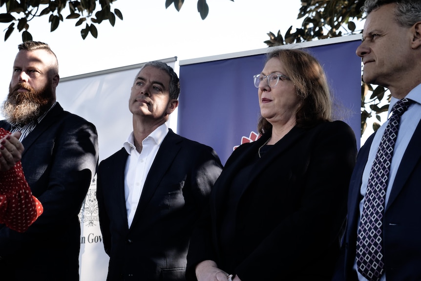 Three men, one with long beard, all wearing suits, an older woman, glasses, wearing black stand together listening and watching.