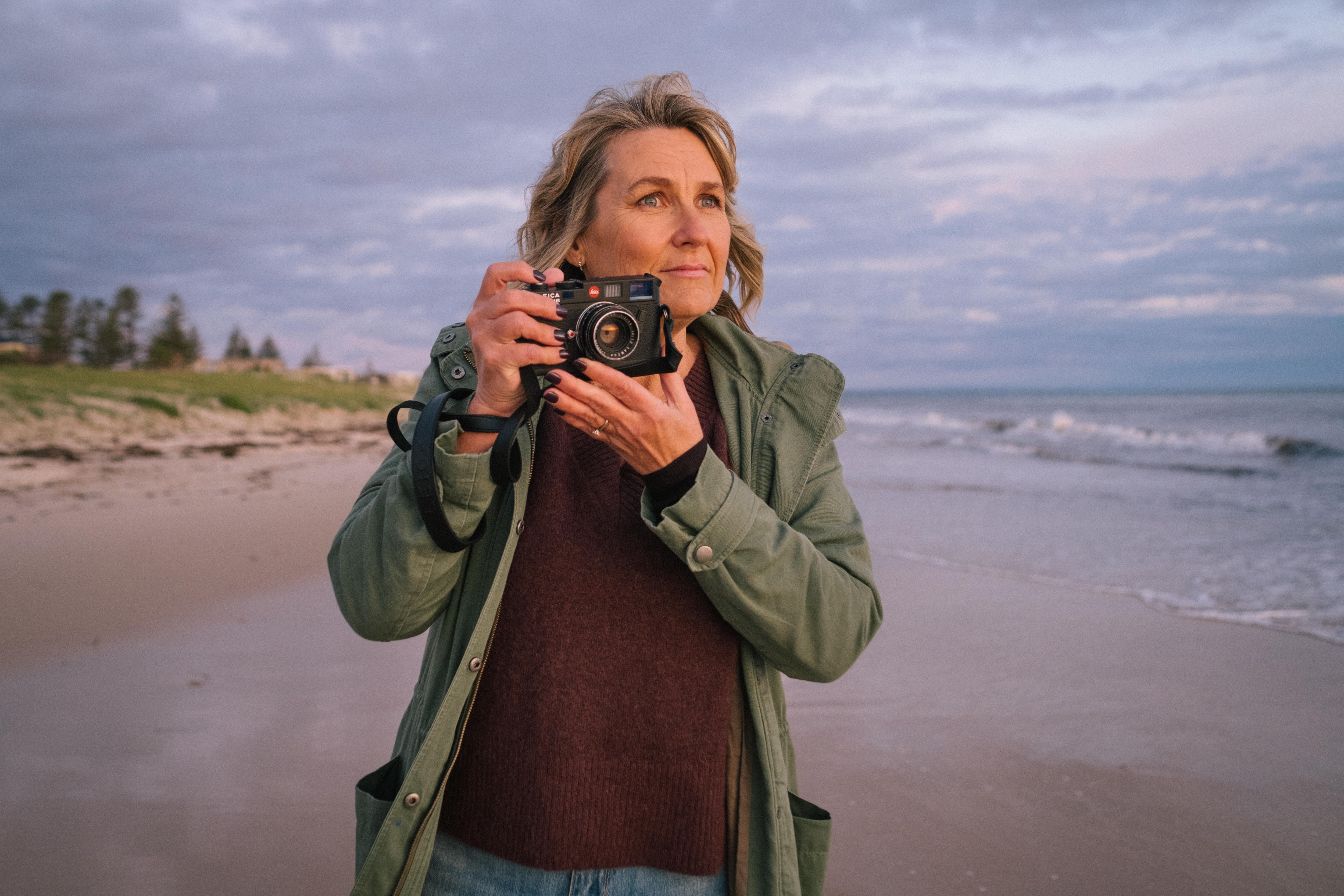 Photographer Narelle Autio stands on a beach on a moody winter's day, holding her camera near her face ready to photograph